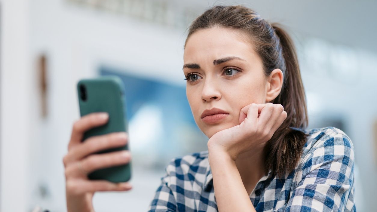 Close up of a depressed Caucasian young woman sitting at home, reading some bad news on her smart phone, feeling sad and worried