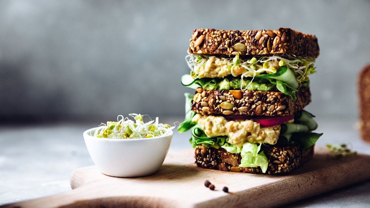 Vegetarian sandwich made with sourdough bread, avocado creme, cucumber, radish and remoulade sauce with bowl of sprouts served on a table.