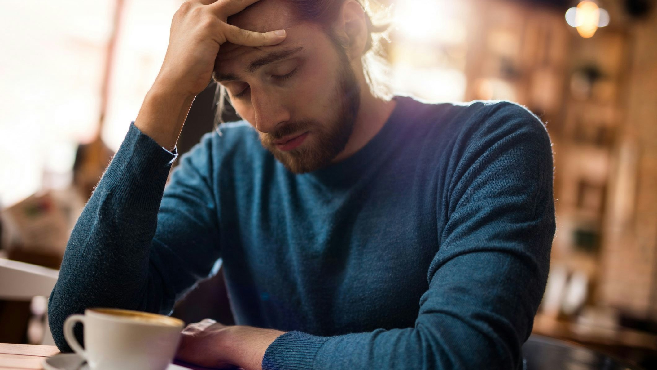 Young man with eyes closed having a headache wxxxhile sitting in a cafe and holding his head in pain.