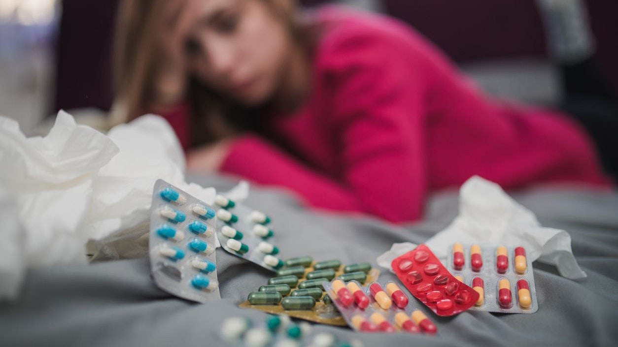 One woman, lying on bed in bedroom, surrounded by pills because she is sick.