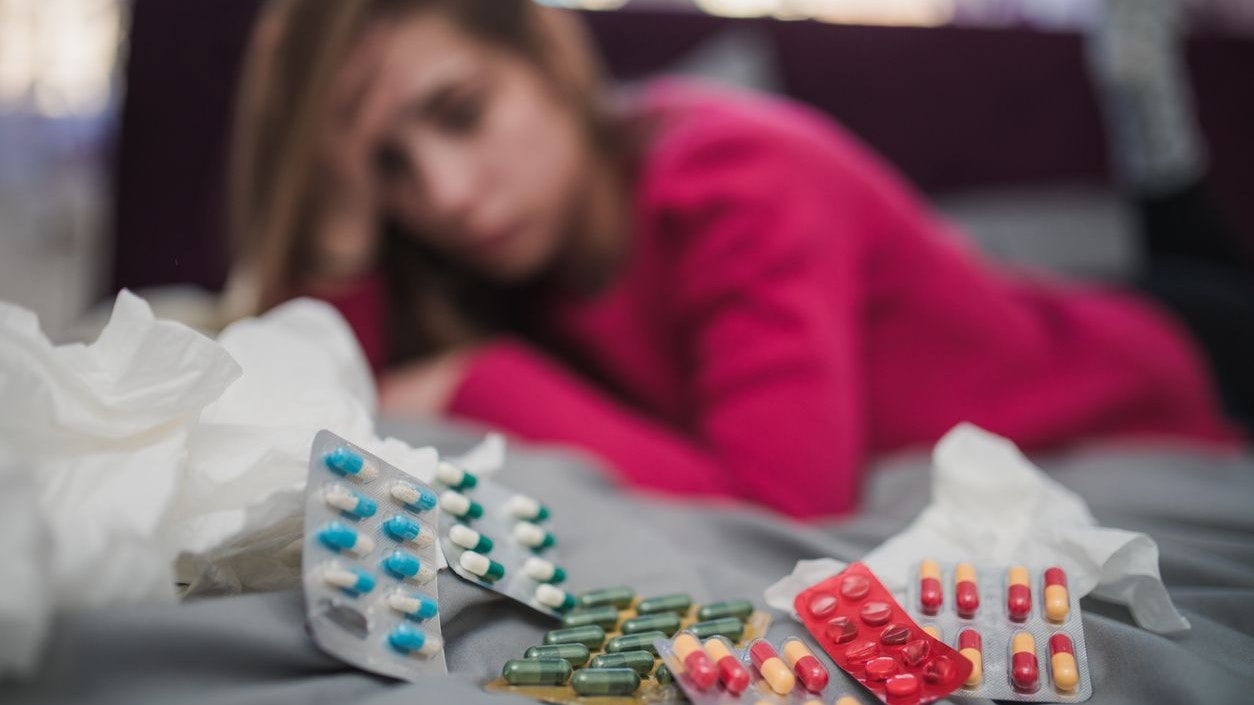 One woman, lying on bed in bedroom, surrounded by pills because she is sick.