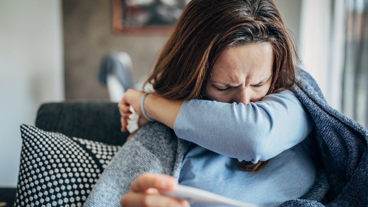One woman, young woman with Covid-19 symptoms sitting on sofa at home, holding thermometer.