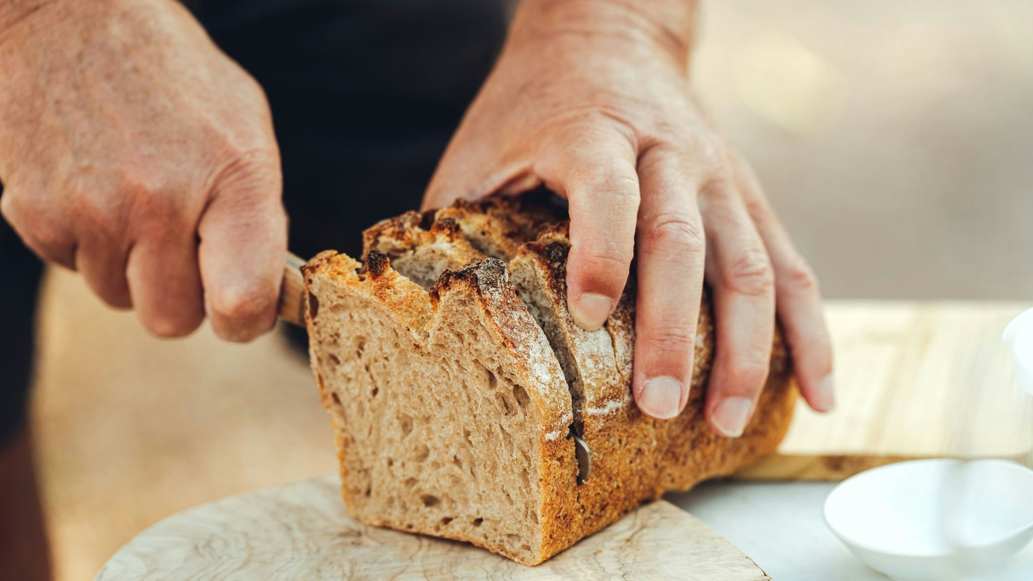 Close-up of senior man hand cutting slice from loaf of bread with kitchen knife in table in garden. male hands  cutting bread on table outdoors.