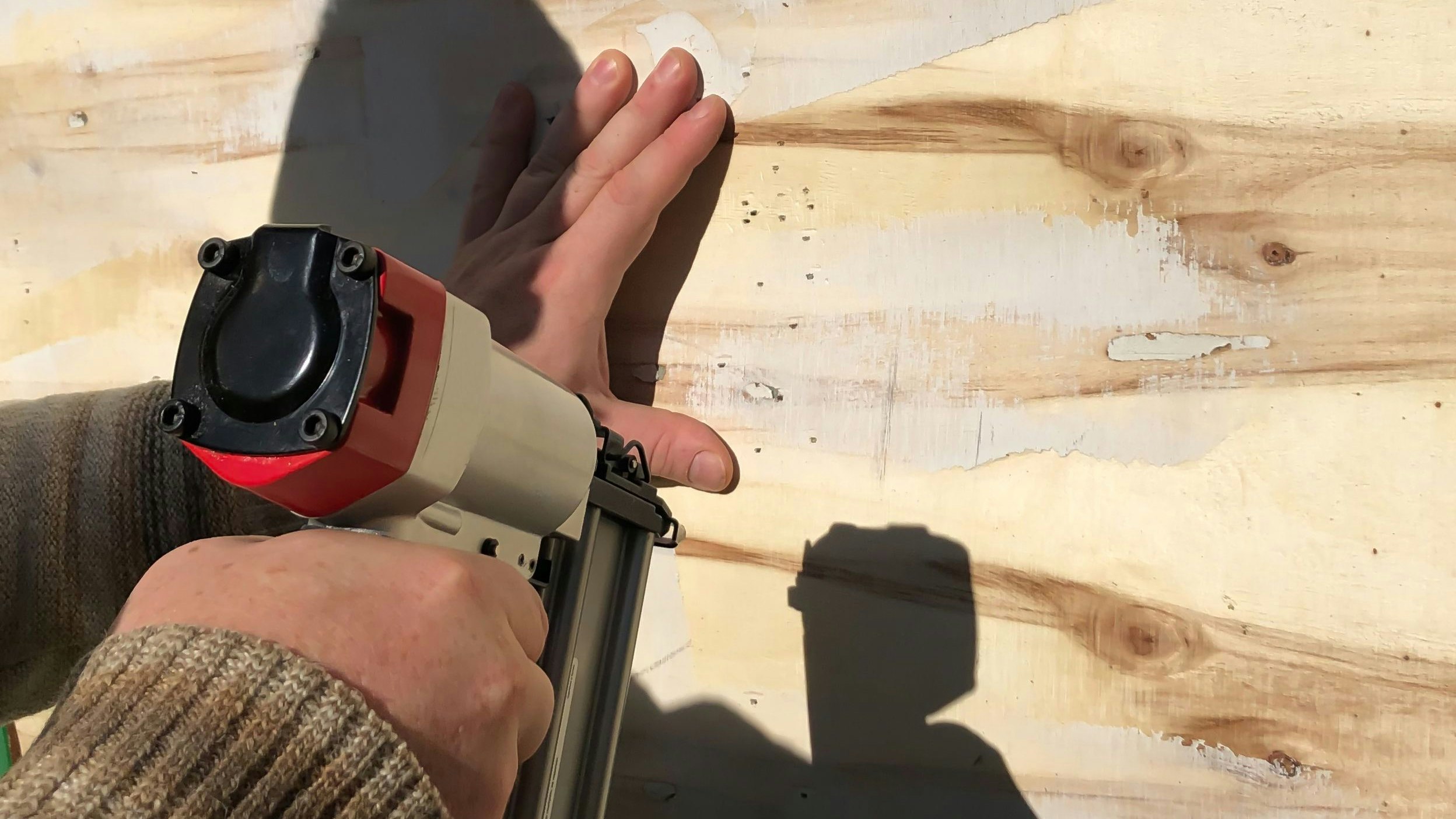 Image of the hands of a man who uses a pneumatic nailer to drive nails into plywood.