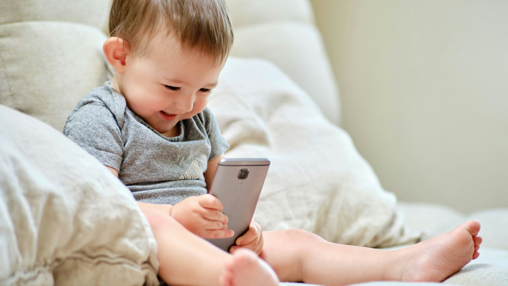 Happy toddler baby boy is sitting with a phone on the sofa in the living room. Child with a smartphone in his hands on the bed