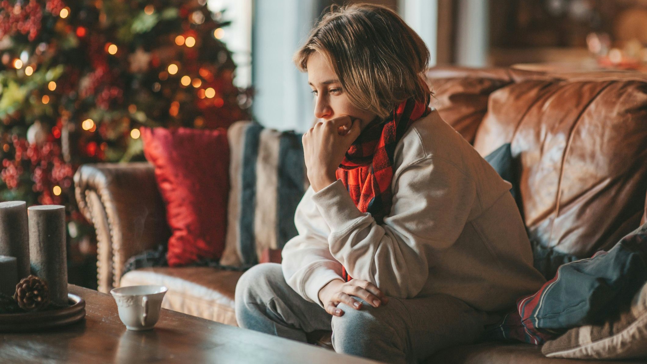 Young teen boy with long hair thoughtful look sad eyes negative mood angry and crying at home. Stylish zoomer gen Z pensive on new year holidays with xmas tree bokeh lights garlands eve 25 december