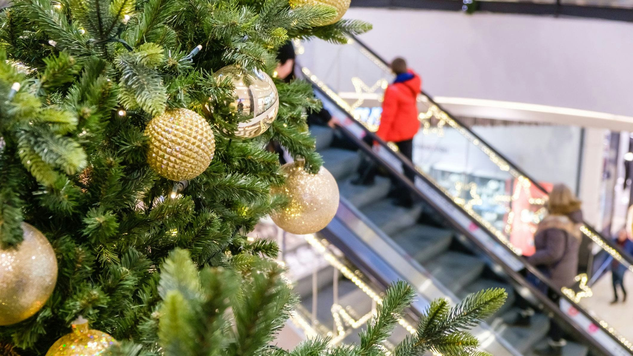 Golden Christmas balls and garland on a decorated fir tree in Shopping Mall. Blurred people on escalator, in festive business center make New Year's purchases,.