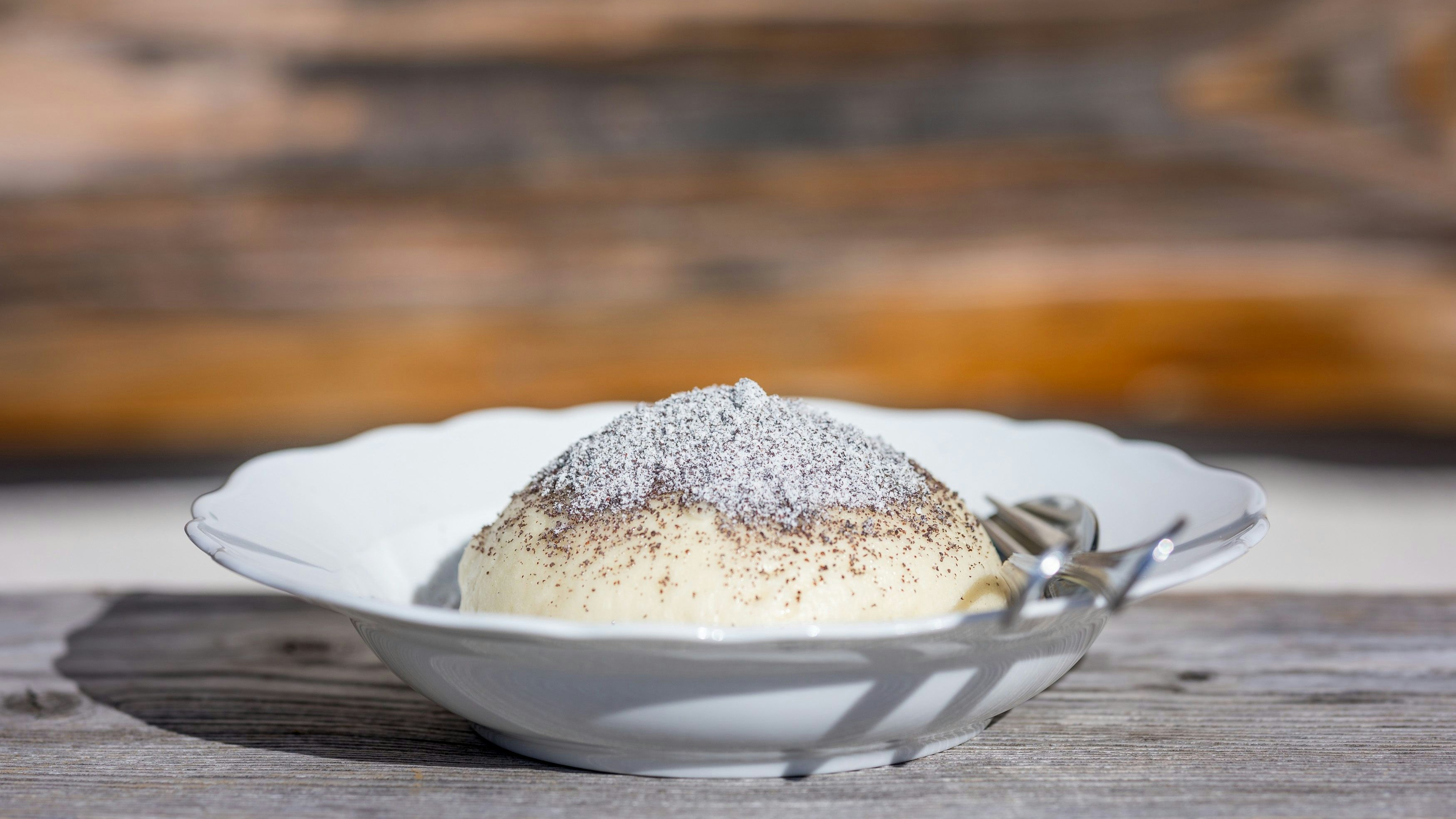 Download von www.picturedesk.com am 27.01.2024 (23:22).  09.02.2018, Gröbming, Steiermark, Österreich (Austria): Ein mit gemahlenem Mohn und Staubzucker bestreuter Germknödel in einem weißen Teller, der samt Besteck auf einem Holztisch vor einer Holzwand einer Alm steht. Der Germknödel gehört zu den beliebtesten Süßspeisen in Österreich und ist als Klassiker der österreichischen Küche ein typisches Essen in vielen Skihütten. .Fotocredit: Martin Huber - 20180209_PD13681 - Rechteinfo: Rights Managed (RM)