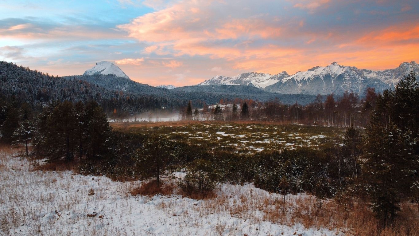 A frozen lake covered in fog is in focus surrounded by forests.  In the far distance you can see the snow-capped mountains of the Alps, hiding the setting sun, giving the clouds in the sky a reddish-orange color.