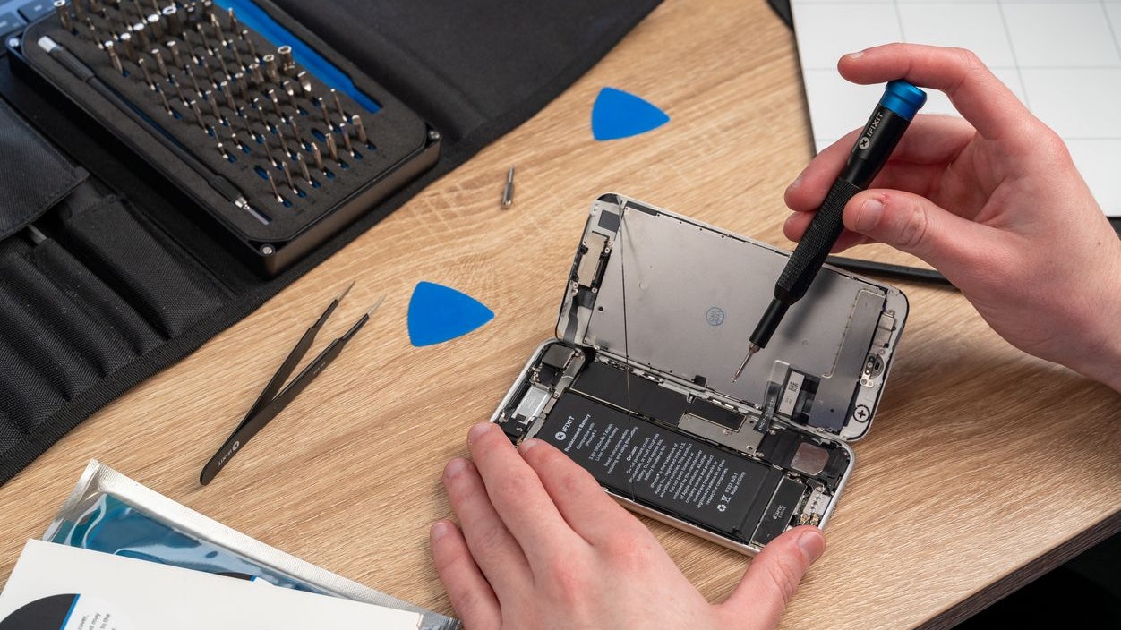 A man performs a self repair on an older iPhone to extend its life, using an iFixit Pro Tech Toolkit and a magnetic project mat. Several opening picks, a pair of precision tweezers, and a precision bit driver is used.
