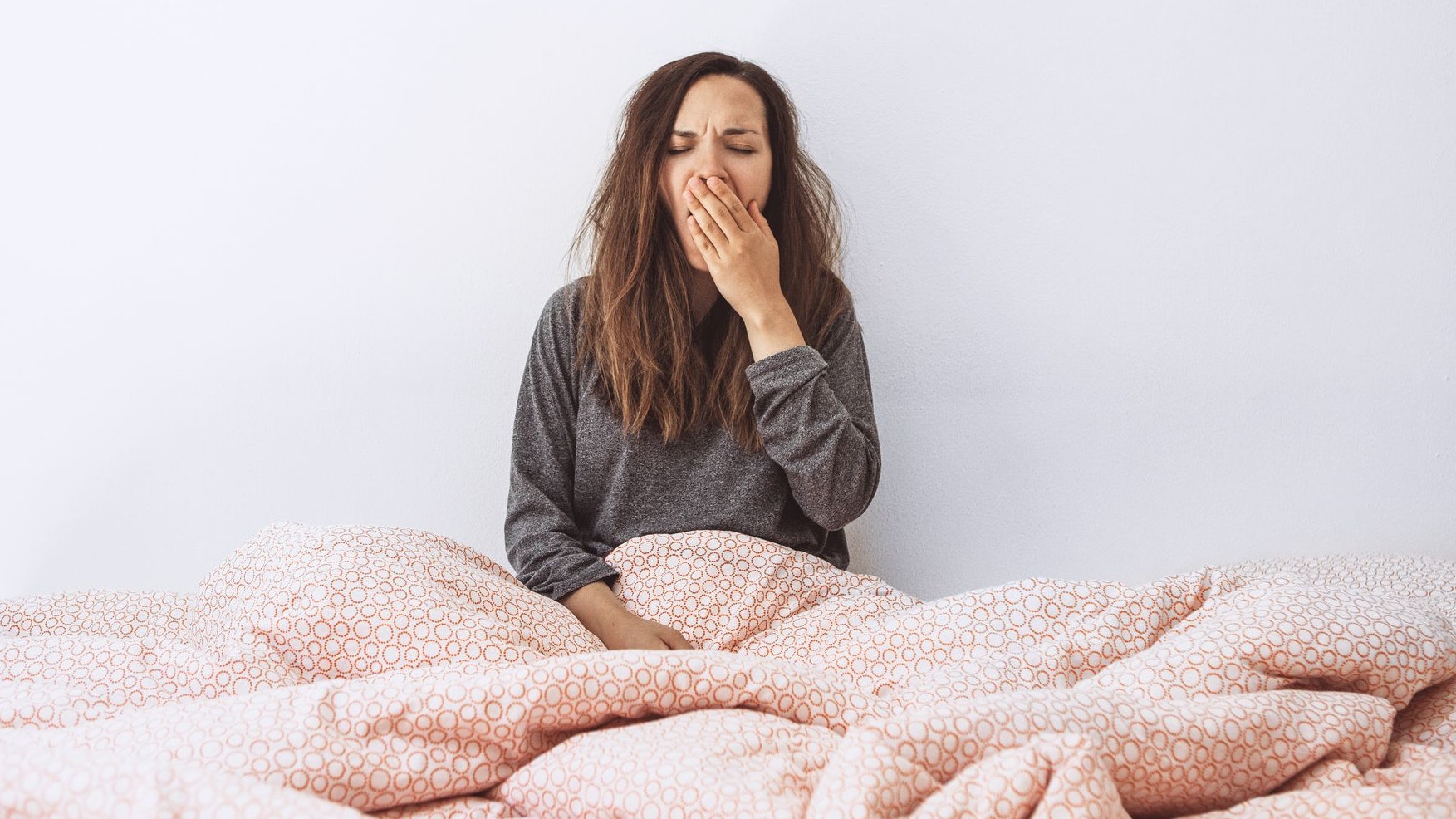 The girl is in bed and yawns. She covers her mouth with her hand. Early morning and she is sleepy.