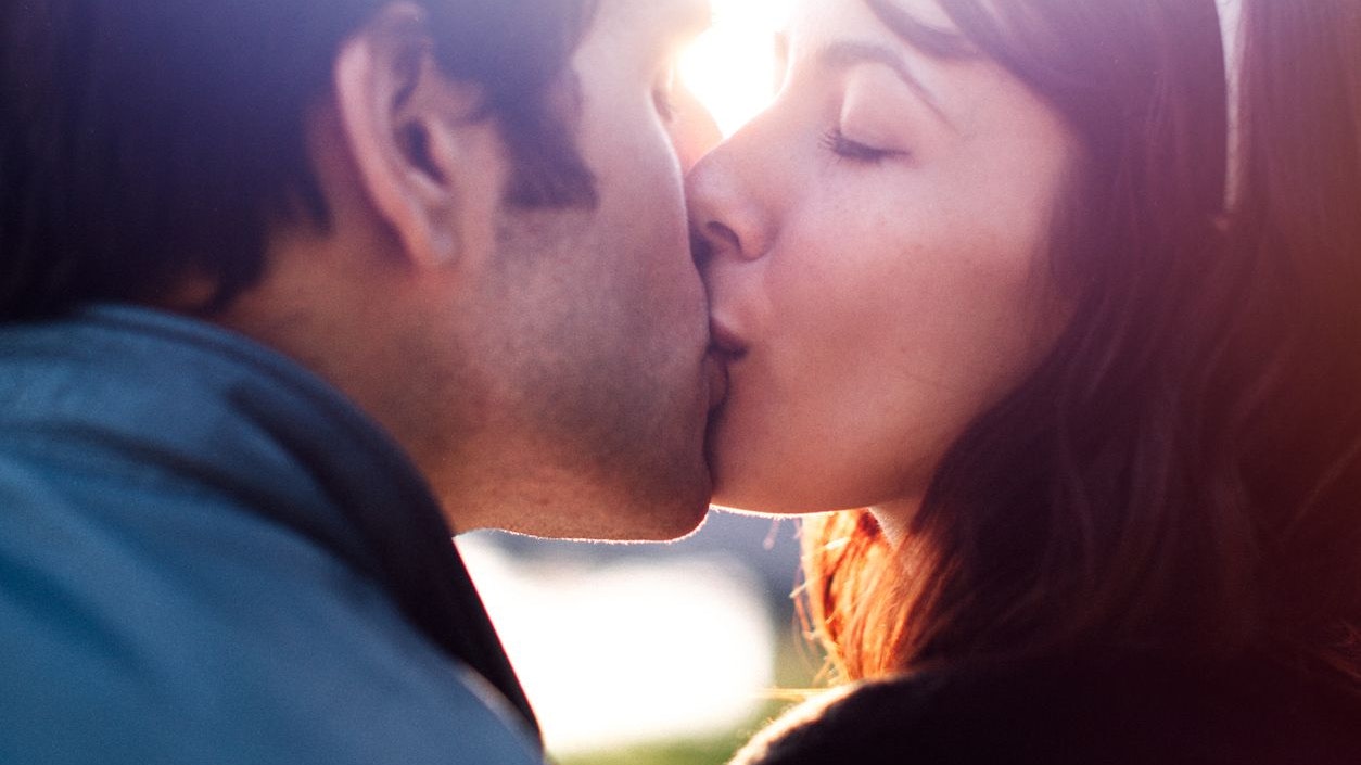 A close up of an attractive young adult couple enjoying a kiss during a romantic date outdoors, the sun shining through with a flare between their faces.