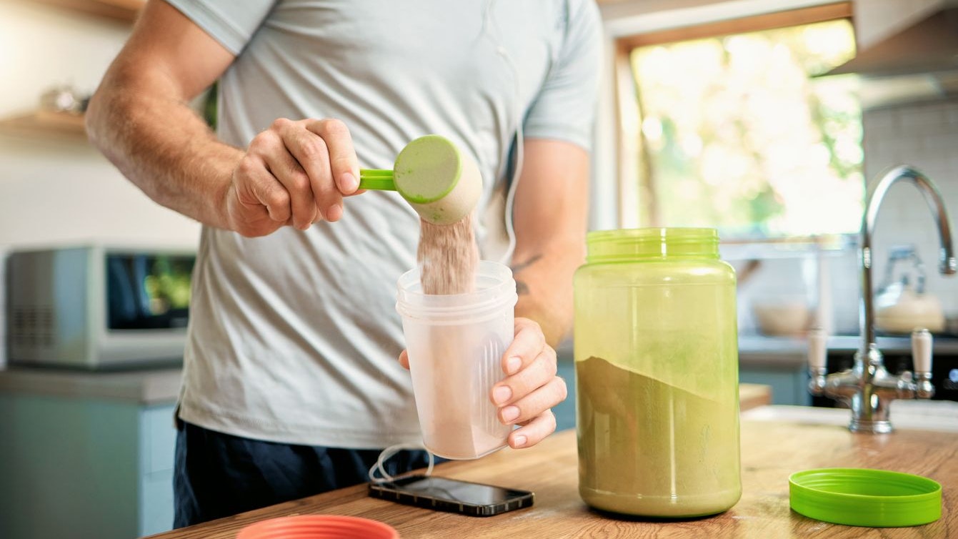 Closeup of one caucasian man pouring a scoop of chocolate whey protein powder to a health shake for energy for training workout in a kitchen at home. Guy having nutritional sports supplement for muscle gain and dieting with weightloss meal replacement