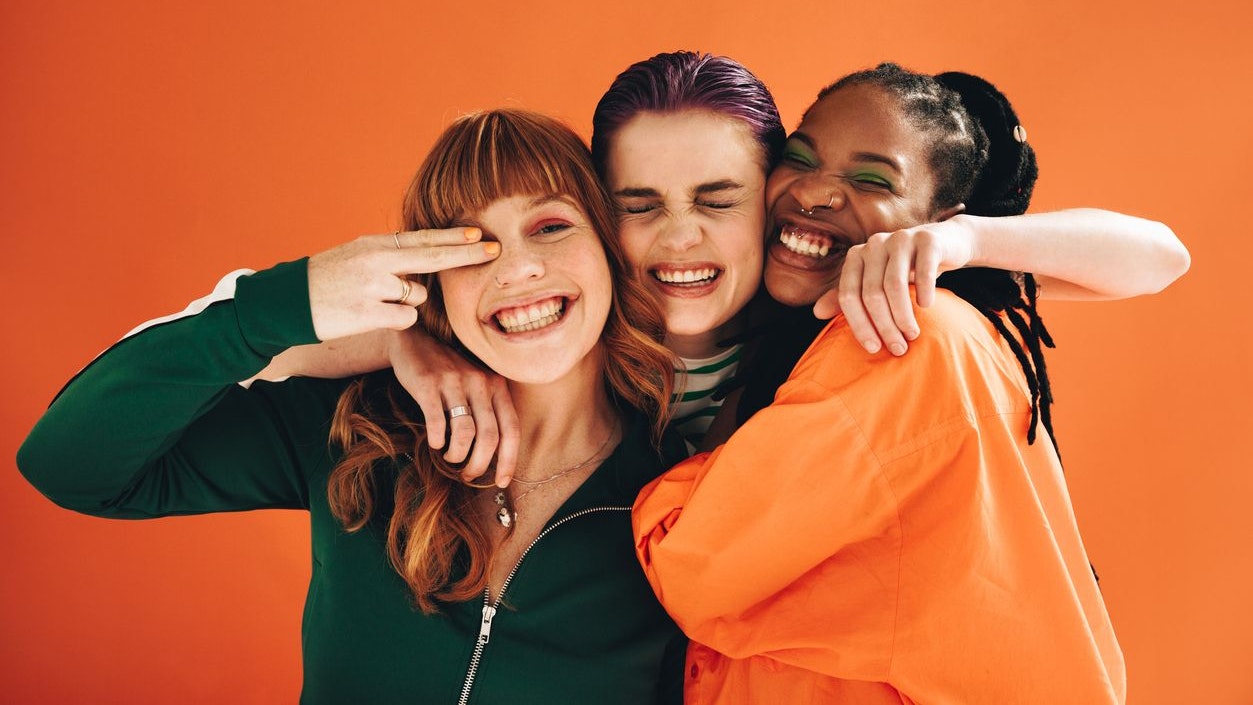 Three multicultural female friends smiling and embracing each other in a studio. Group of vibrant young women enjoying themselves while standing against an orange background.