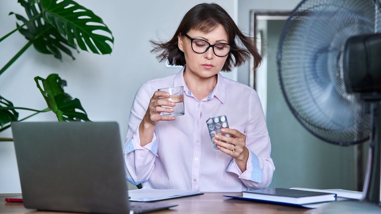 Mature woman with glass of water blister of pills taking medicine vitamins to relieve menopause symptoms, sitting at desk at workplace with computer laptop