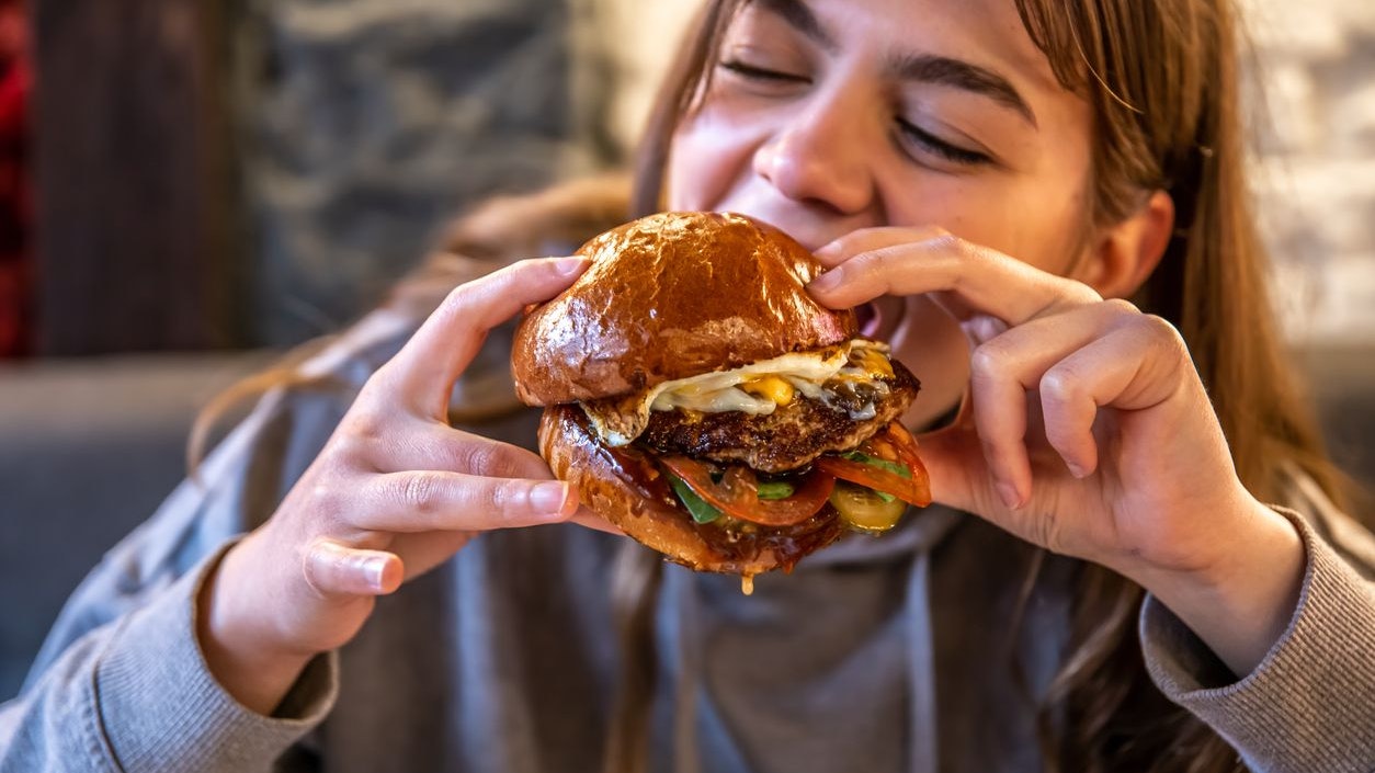 Close-up of a girl eating a big burger with meat and vegetables on a blurred background.