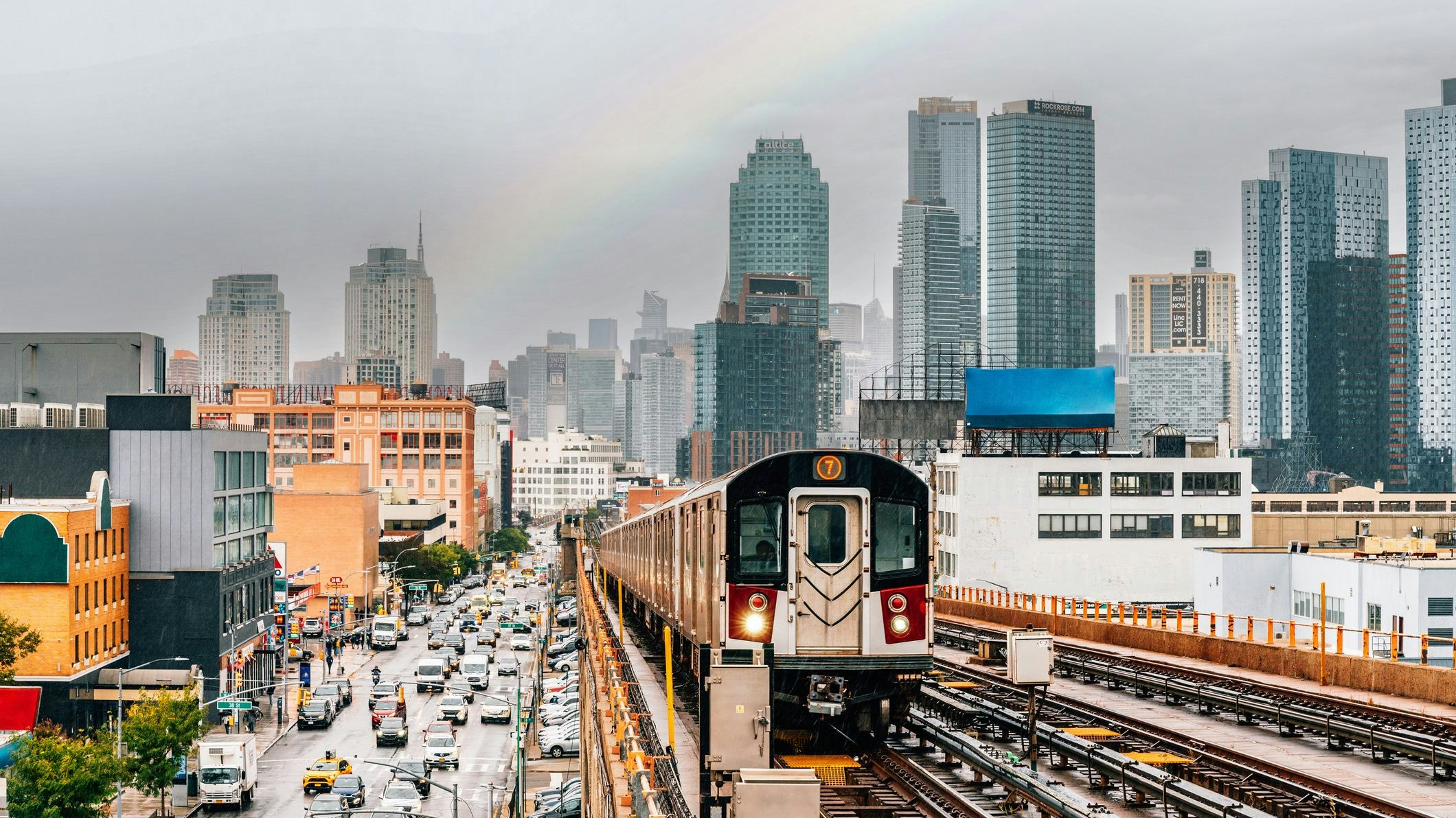 New York City subway train is approaching an elevated subway station in Queens. Raining day in New York. Manhattan skyline in the background.