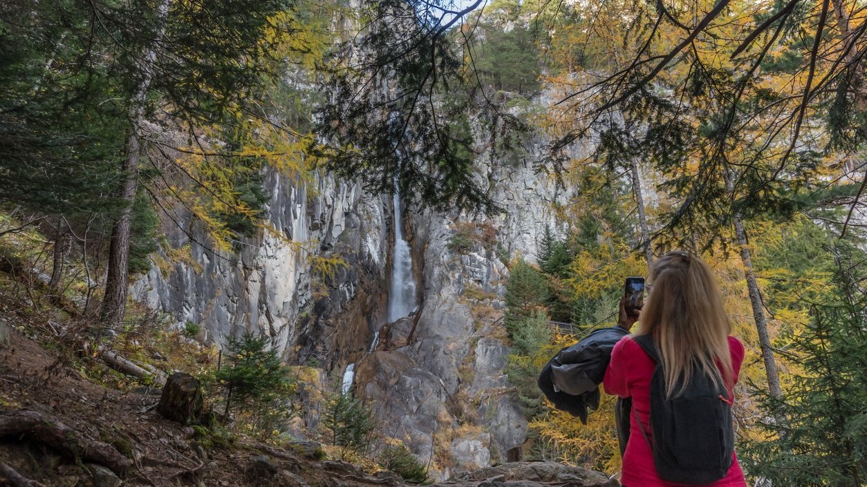 Woman with mobile phone taking photos of the waterfall on a rocky mountain in a forest. Nature, travel and technology concept.