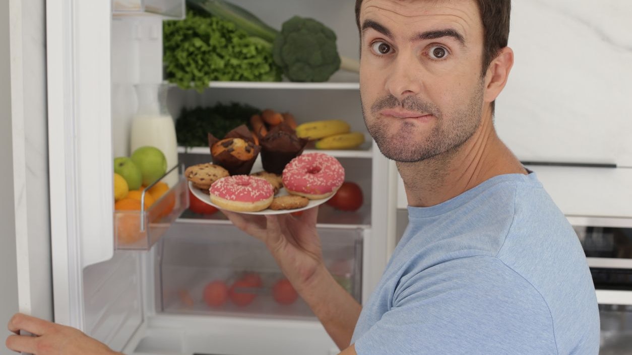 An uncertain looking man is choosing junk food over vegetables. He holds a plate with muffins and doughnuts in front of a refrigator fill with healthy food.