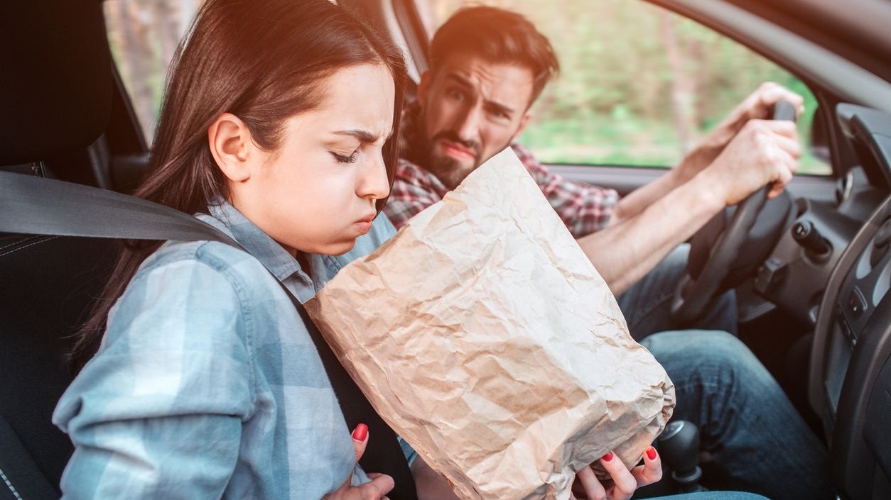 A picture of sick girl holding a bag and trying to vomit into it. She feels bad. Girl is holding her hand on stomach. Guy is looking at her with sight full of disgution