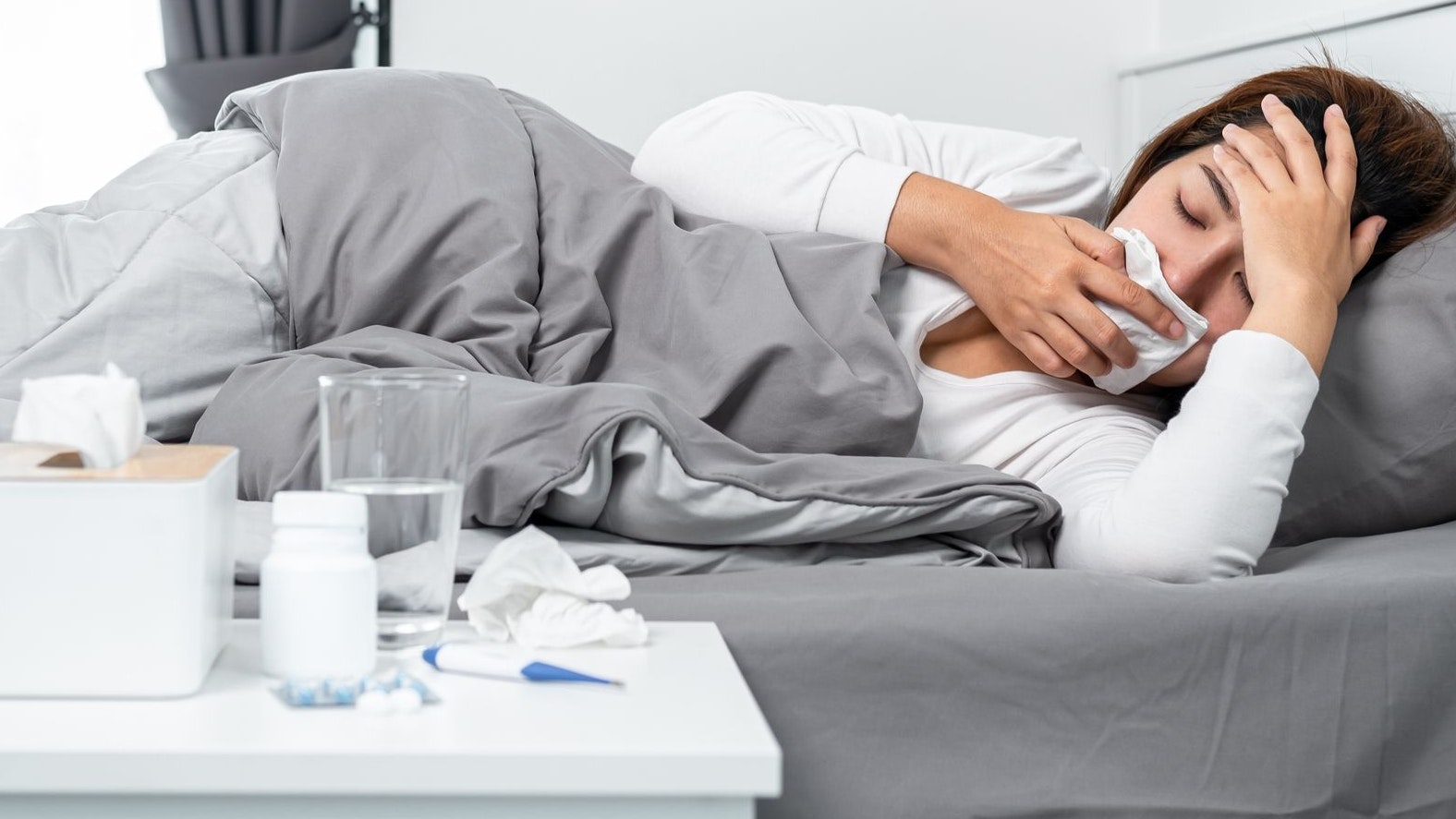 Asian woman using tissue to cough, sneezing and touching forehead while feeling sick and lying in blanket on the bed after using thermometer to checking body temperature and take medicine in bedroom