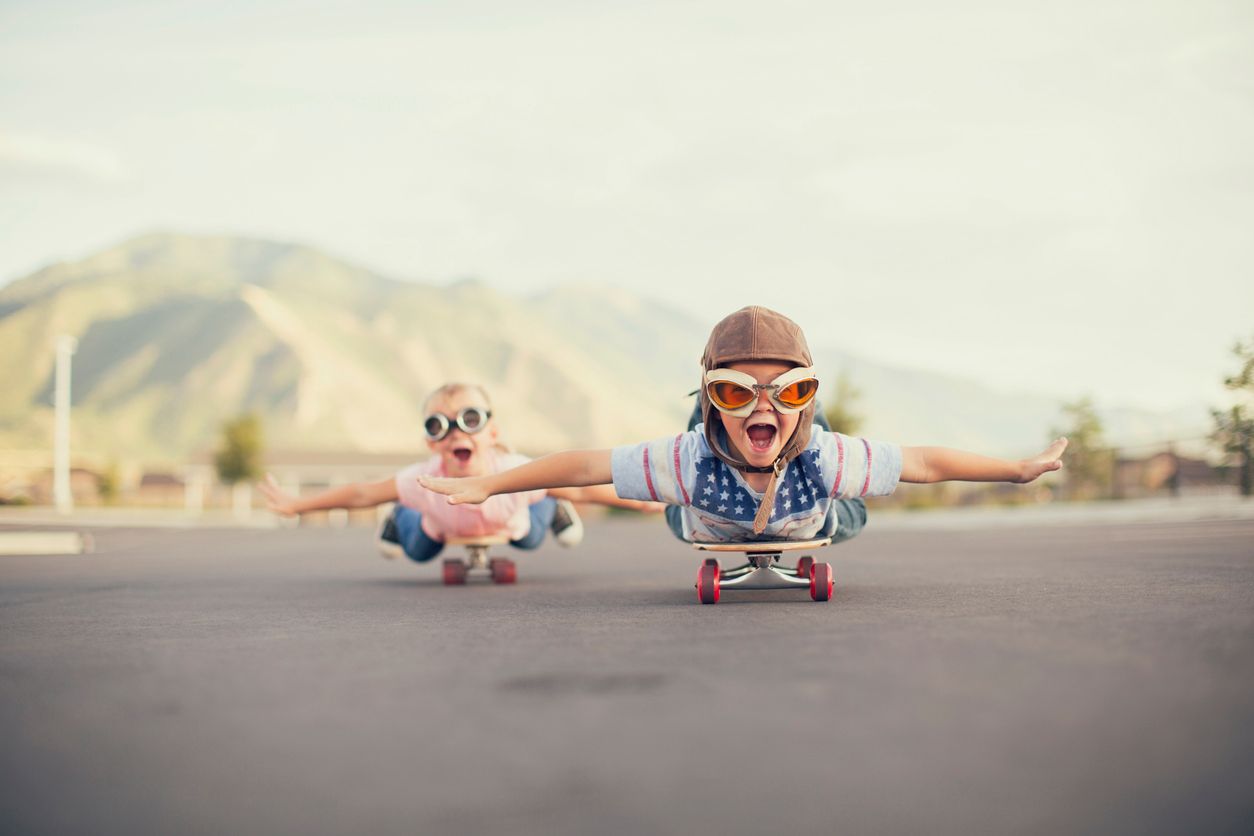 A young boy and girl are wearing flying goggles while outstretching their arms to attempt flying while on skateboards. They have large smiles and are imagining taking off into the sky.
