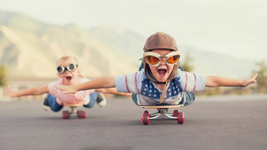 A young boy and girl are wearing flying goggles while outstretching their arms to attempt flying while on skateboards. They have large smiles and are imagining taking off into the sky.