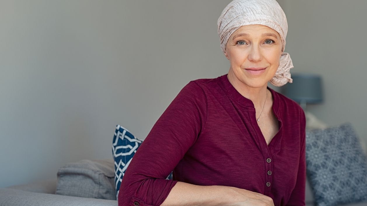 Mature woman with cancer in pink headscarf smiling sitting on couch at home. Smiling woman suffering from cancer sitting after taking chemotherapy sessions. Portrait of mature lady facing side-effects of hair loss, copy space.
