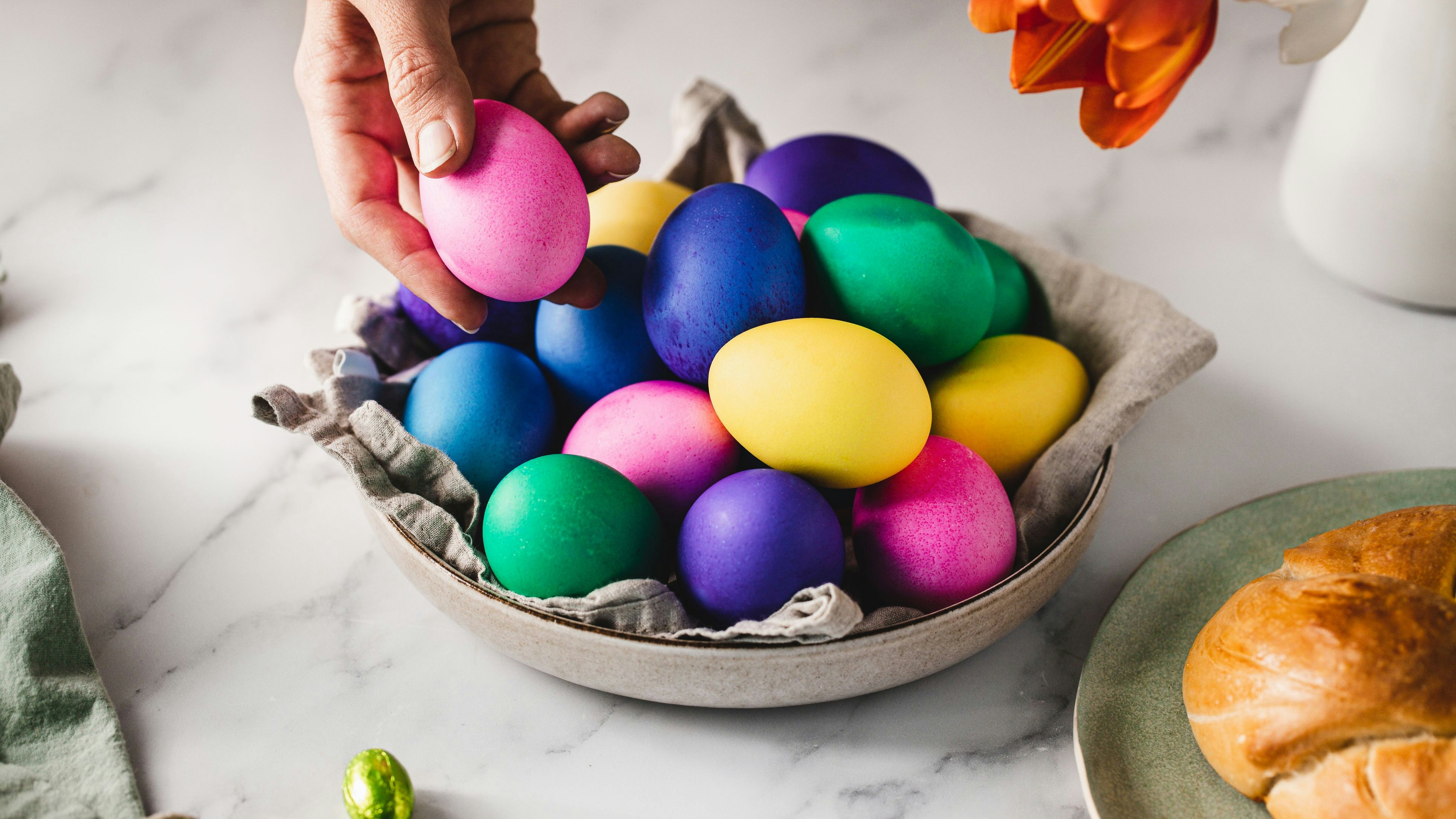 Close-up of a woman hand placing pink colored egg in a bowl full of Easter eggs over breakfast table. Woman decorating breakfast table for Easter.