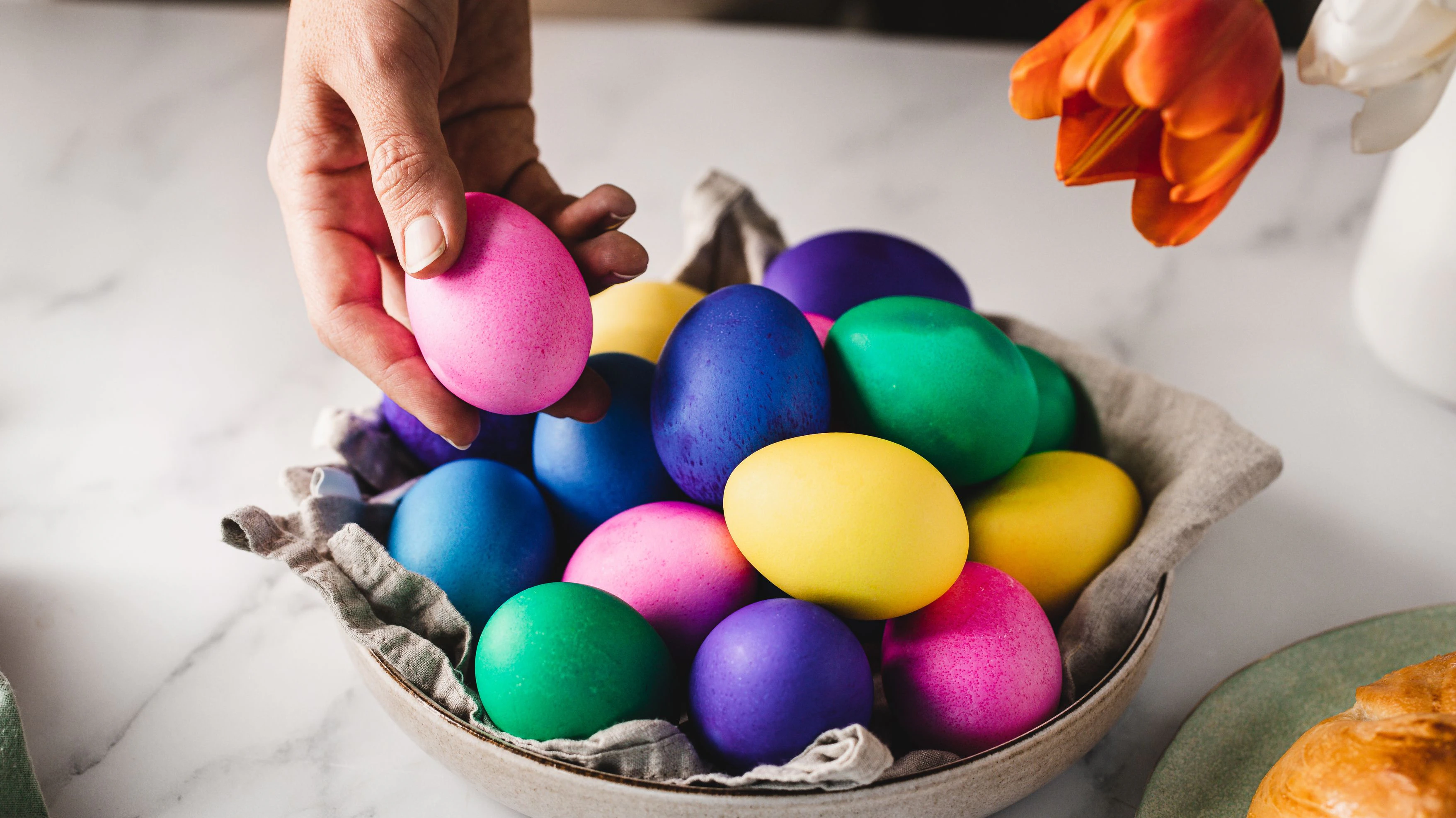 Close-up of a woman hand placing pink colored egg in a bowl full of Easter eggs over breakfast table. Woman decorating breakfast table for Easter.