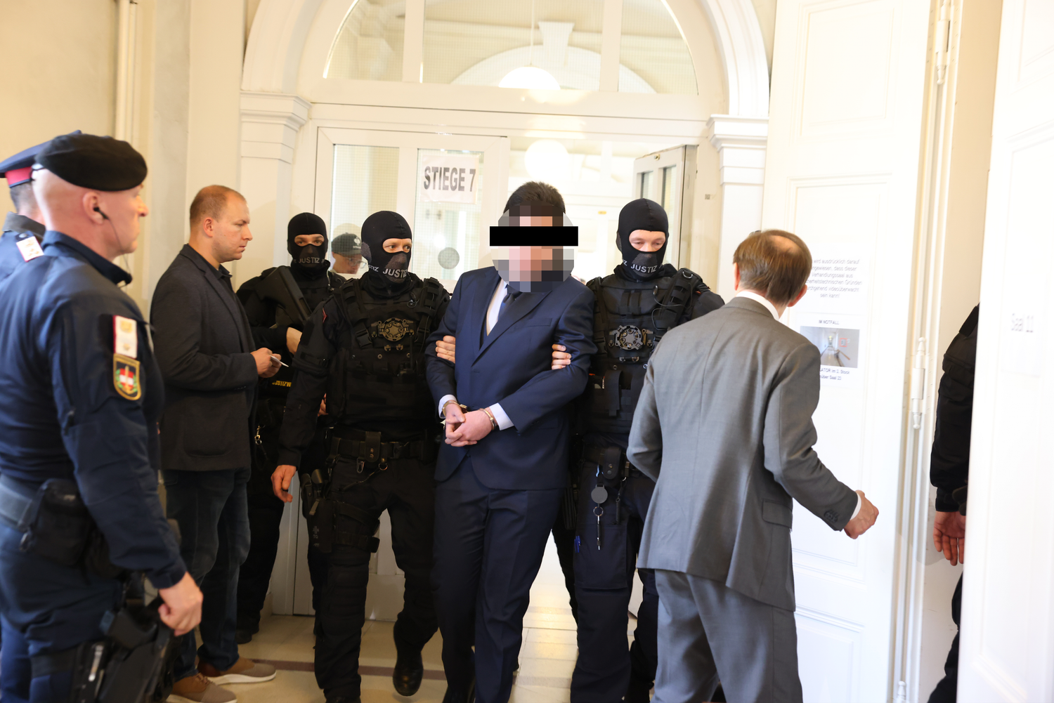 In seinem dunkelblauen Sonntagsanzug wird der Angeklagte von schwerbewaffneten Justizbeamten vorgeführt.