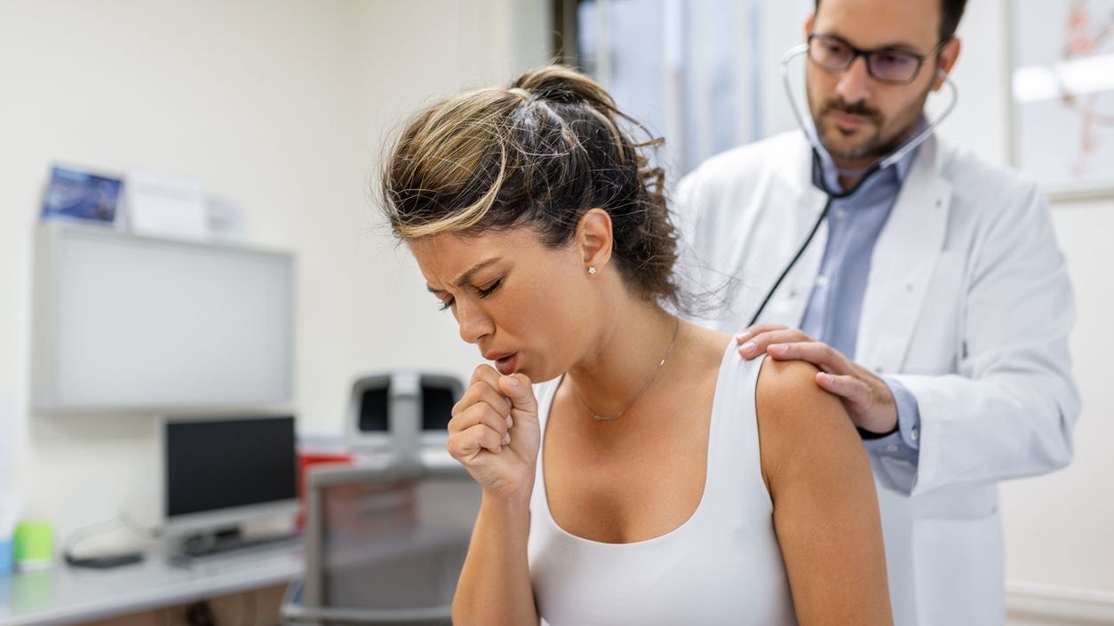Young female patient in the clinic suffered from pneumonia, she is coughing the doctor listens to the wheezing in the lungs with a stethoscope.