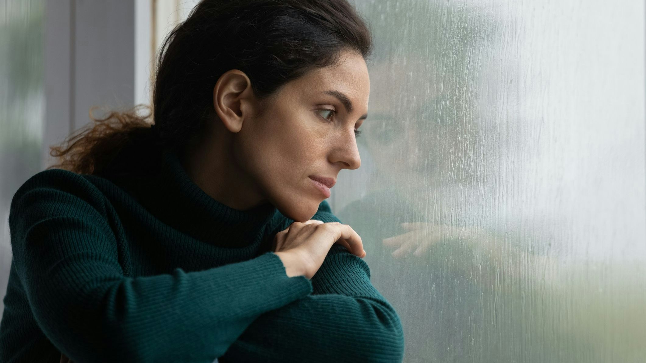 Thoughtful stressed young hispanic latin woman sitting on windowsill, looking outside on rainy weather, having depressive or melancholic mood, suffering from negative thoughts alone at home.