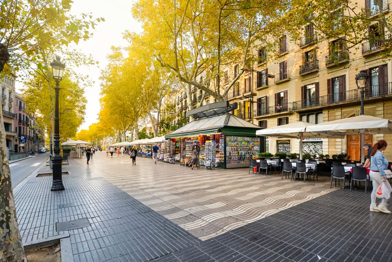 Early morning on the pedestrian Las Ramblas boulevard through historic Barcelona Spain as tourists and locals pass by kiosks, shops, cafes and news stands.