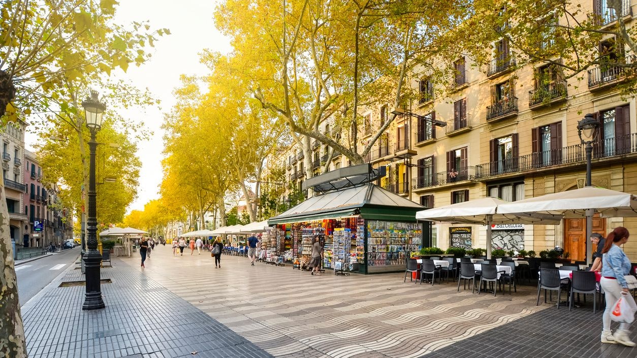 Early morning on the pedestrian Las Ramblas boulevard through historic Barcelona Spain as tourists and locals pass by kiosks, shops, cafes and news stands.