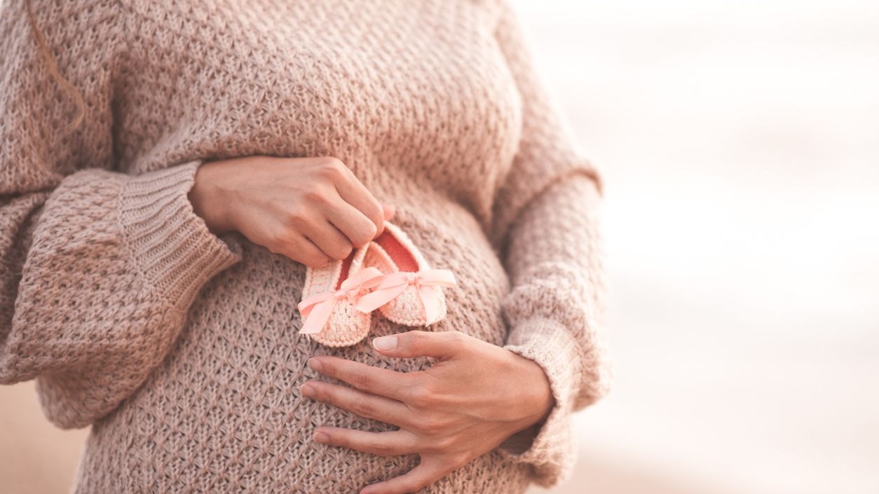 Pregnant woman wearing knitted comfy sweater holding baby shoes outdoors. Motherhood. Maternity. 20s. Healthy lifestyle. Healthcare.