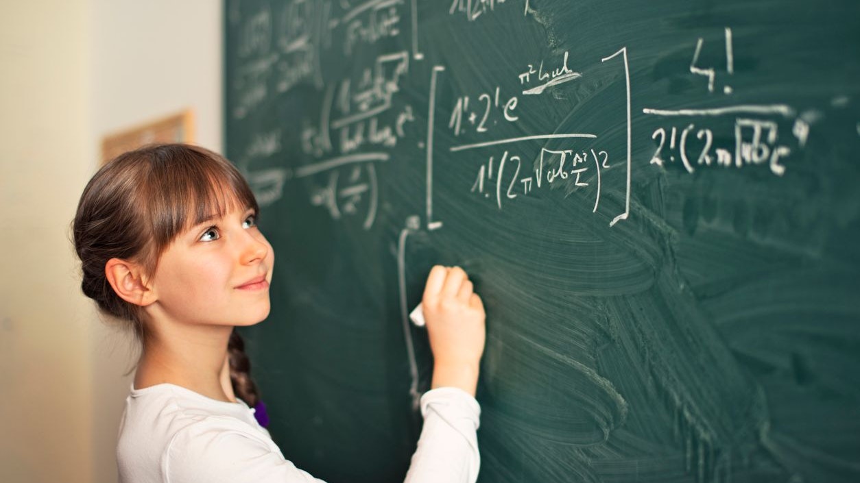 Little mathematics genius girl writing difficult mathematics equations on a green chalkboard. The gilr is smiling and obvioulsy she is having fun solving the equation.