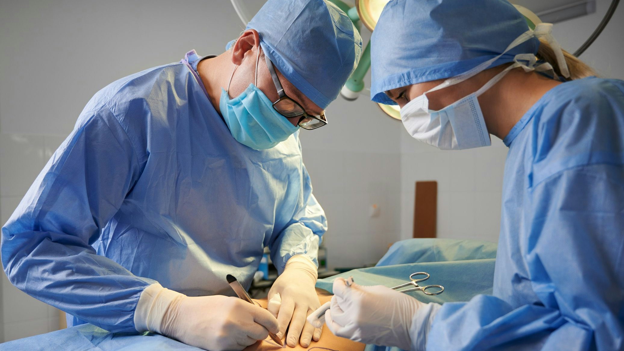 Doctors in surgical hospital suits performing cosmetic surgery in operating room. Medical team wearing protective face masks, sterile gloves and medical caps. Concept of medicine and plastic surgery.