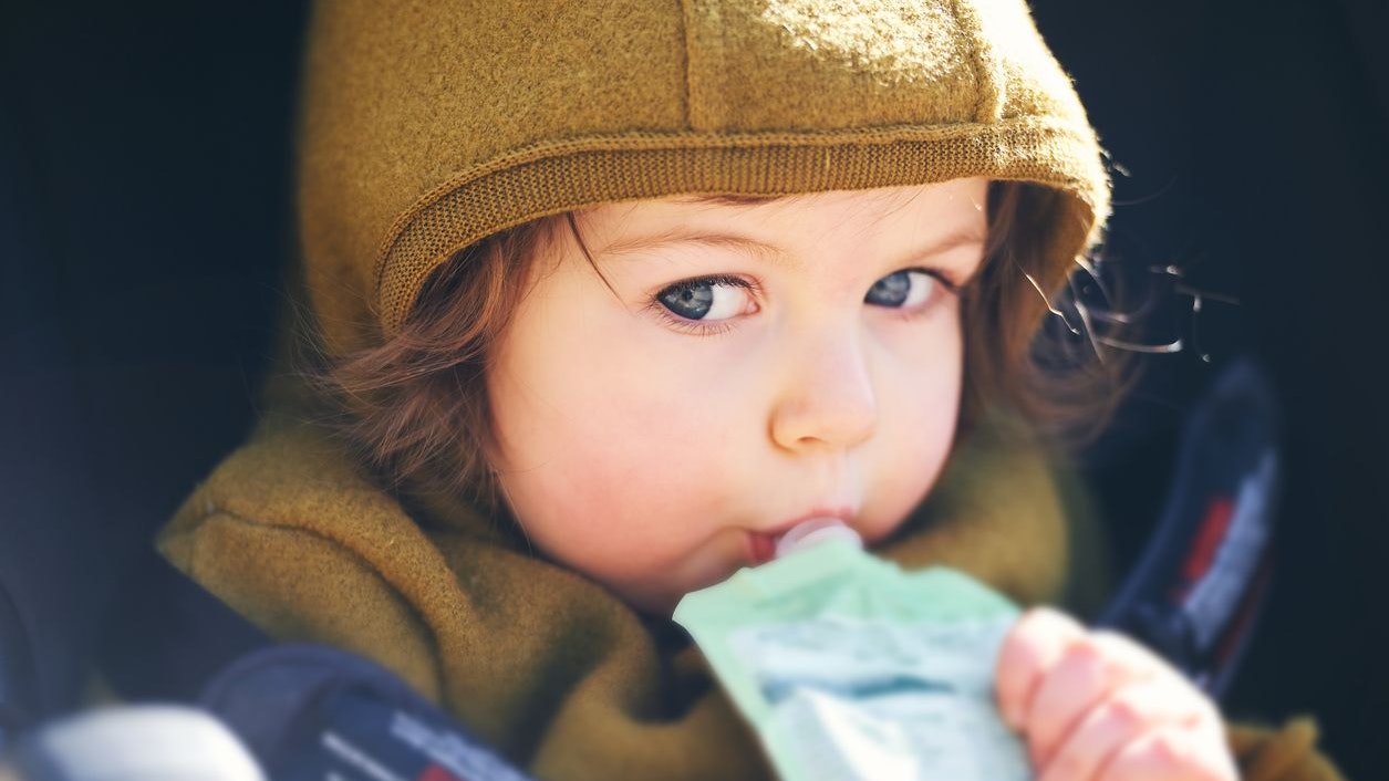 Close up portrait of sweet toddler kid eating fruit puree from plastic doy pack, sitting in stroller, outdoor snack time