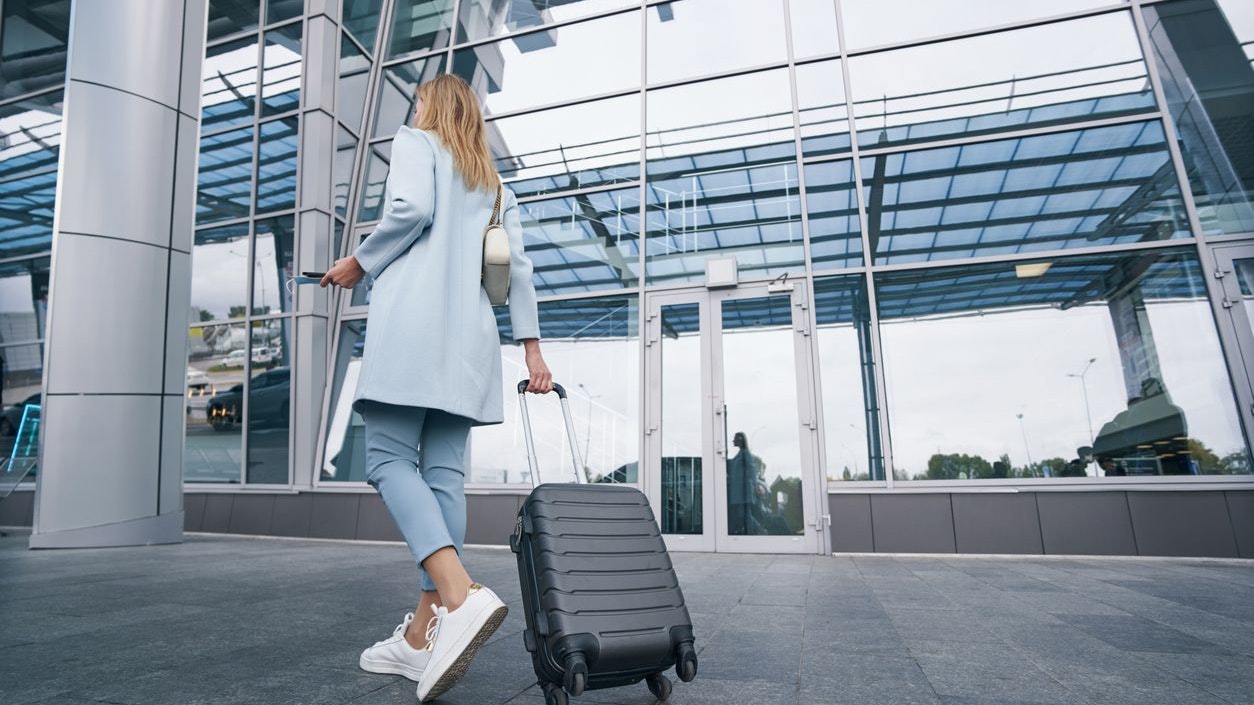 Back view of Caucasian female traveler with luggage standing before airport terminal building