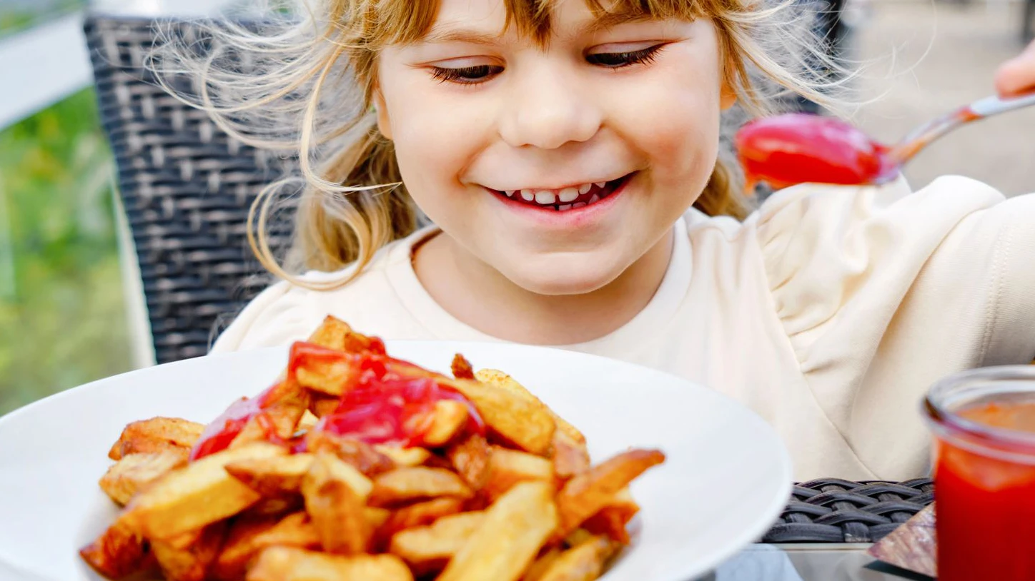 Portrait of happy smiling preschool girl eating french fries with tomato ketchup in restaurant on terrace outdoors. Little child with blond hairs enjoy unhealthy fast food or fresh prepared lunch