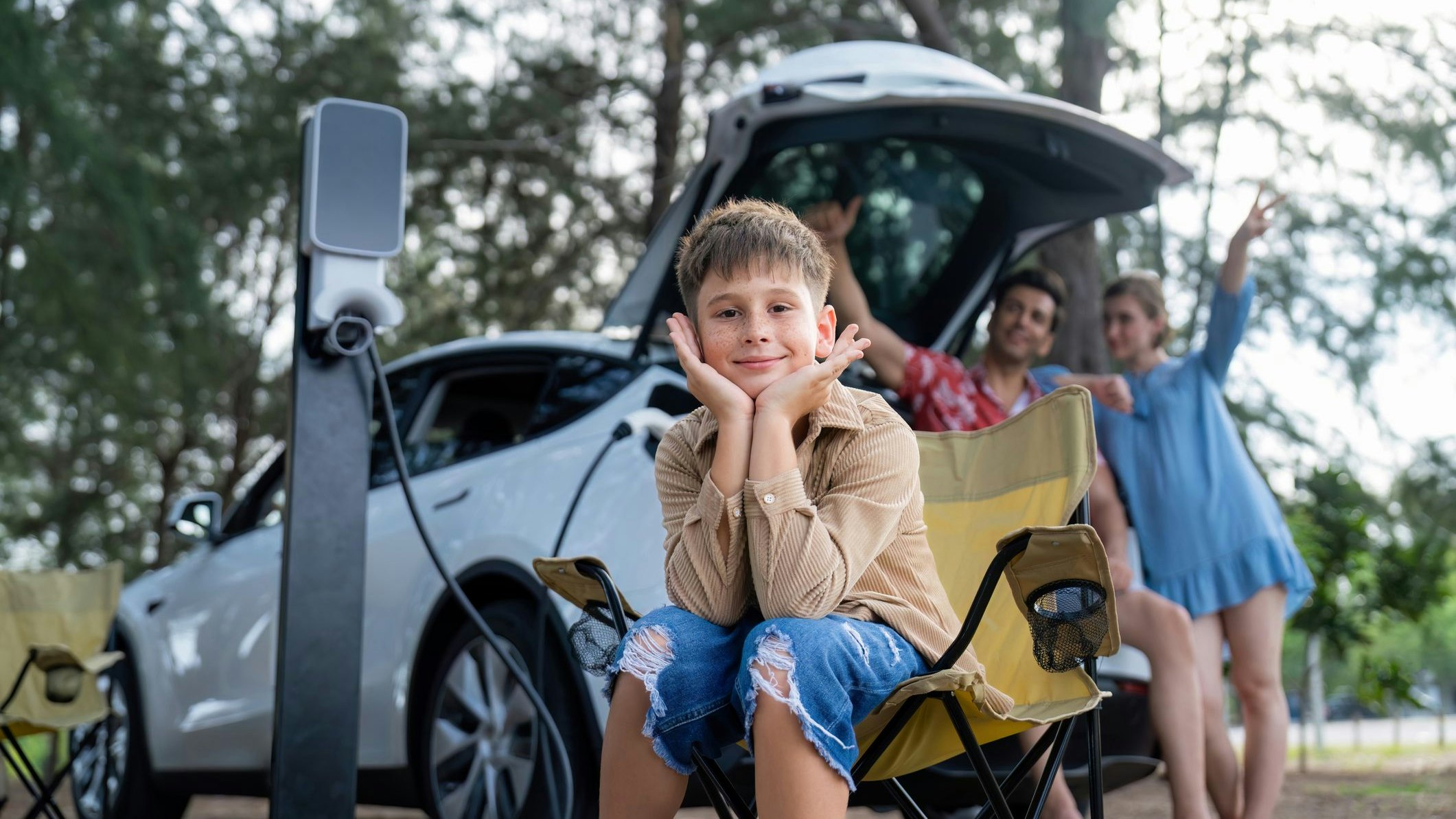 Little boy portrait sitting on camping chair with his family in background. Road trip travel with alternative energy charging station for eco-friendly car concept. Perpetual