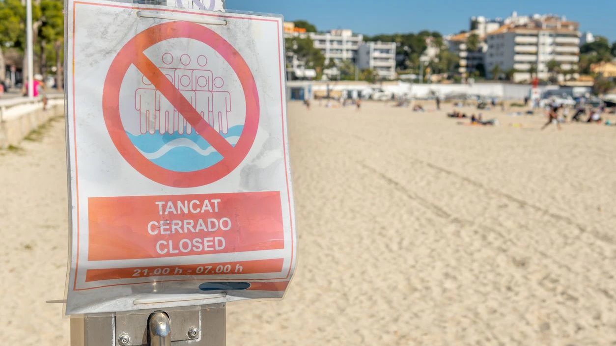 No bathing sign from 21:00 to 07:00 in Spanish, Catalan and English. In the background a beach on the island of Mallorca with tourists and out of focus. New normal concept