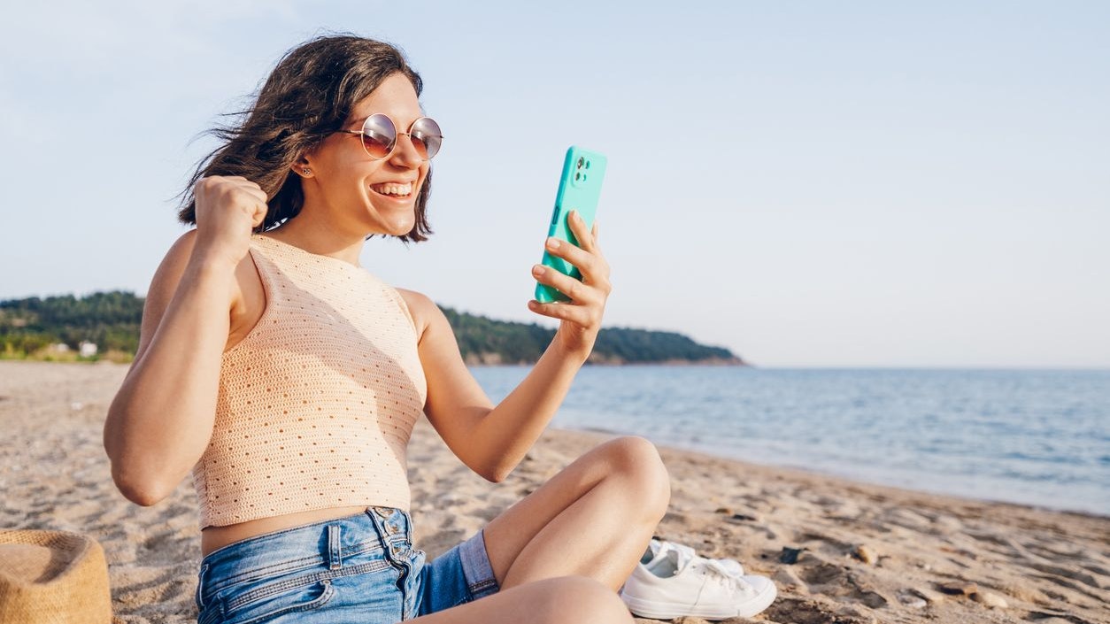 A young woman rejoicing because of the good news she saw on her phone while sitting on the beach.