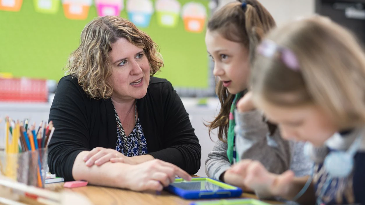 A teacher helps two of her first graders at their desks. They have digital tablets in front of them and she is going over an assignment with them before they start working on it themselves on the tablets.