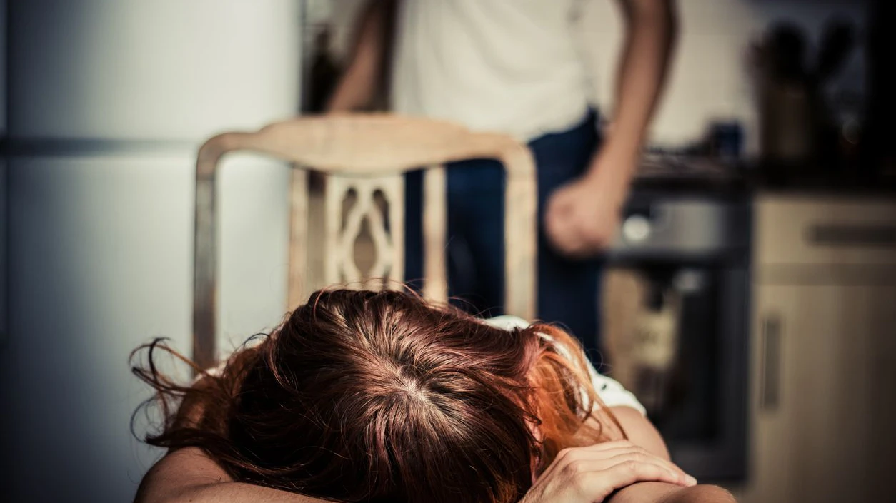 Young woman is reesting her head on a kitchen table, her abusive partner hovering in the background