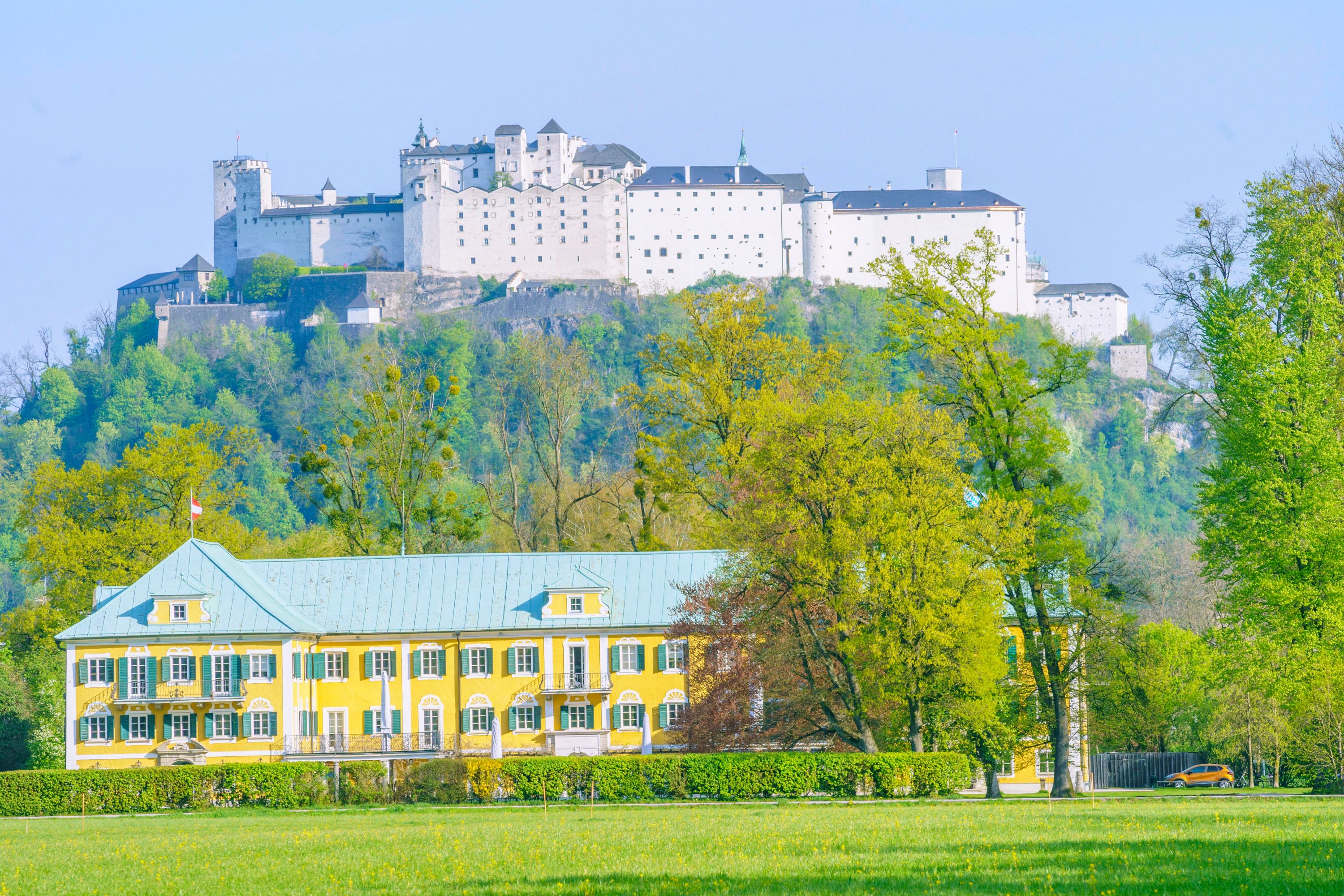 Blick in der Stadt Salzburg entlang der Hellbrunner Alllee über grüne Wiesen zum Gwandhaus und weiter zum Festungsberg mit der Festung Hohensalzburg.