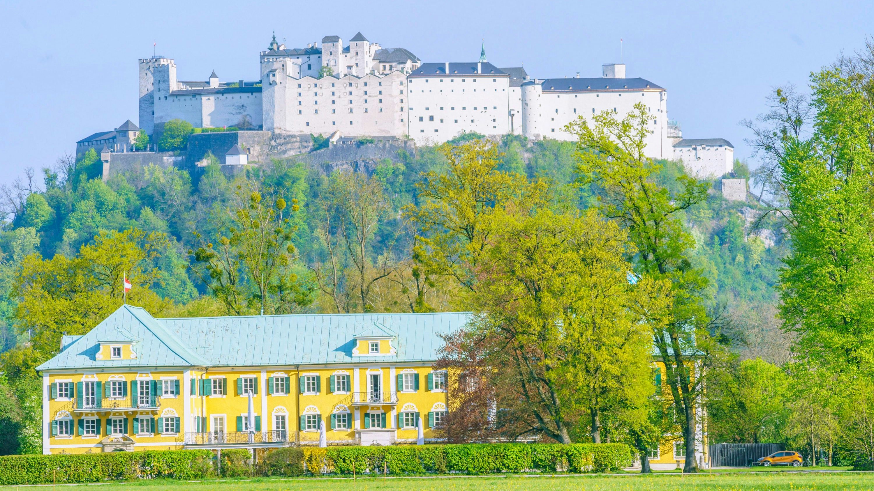 Blick in der Stadt Salzburg entlang der Hellbrunner Alllee über grüne Wiesen zum Gwandhaus und weiter zum Festungsberg mit der Festung Hohensalzburg.