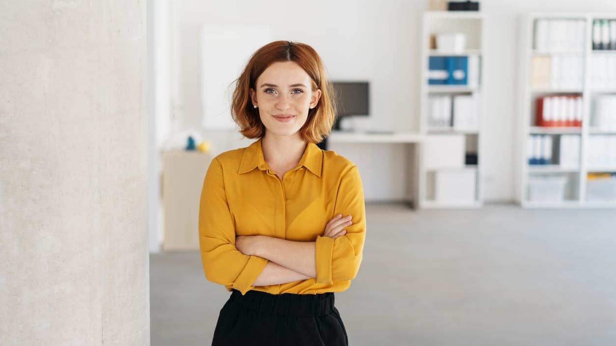 Happy relaxed confident young businesswoman standing with folded arms in a spacious office looking at the camera with a warm friendly smile
