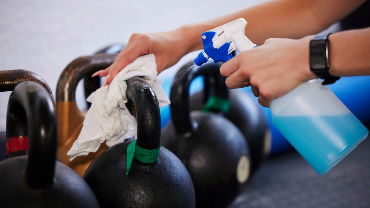 Gym, hygiene and hands cleaning kettlebell with cloth and spray bottle, dirt and germs at fitness studio. Janitor, cleaner or caretaker at sports club washing dust and bacteria from workout equipment
